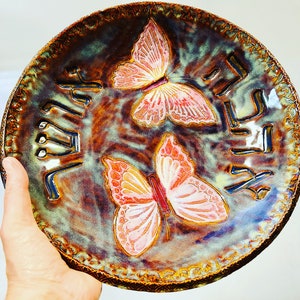 May include: A ceramic plate with a brown, green, and blue glaze. The plate features two pink butterflies with white accents. The plate has a decorative border and the word "Butterfly" written in cursive on the plate.