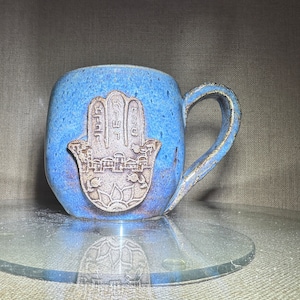May include: A blue ceramic mug with a hamsa hand design. The hamsa is a symbol of protection and good luck.