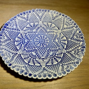 May include: A round, white and blue ceramic plate with a detailed lace-like pattern. The plate has a scalloped edge and a central design that resembles a doily. The blue and white color scheme gives it a classic, elegant look.