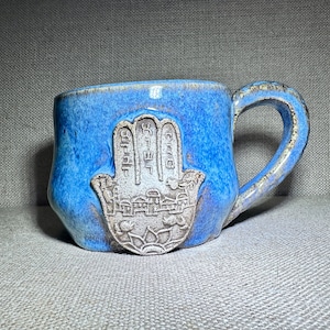 May include: A handmade ceramic mug with a blue glaze. The mug features a raised, detailed design of a Hamsa hand with Hebrew text and a city scene. The handle is textured, and the mug is suitable for hot beverages.