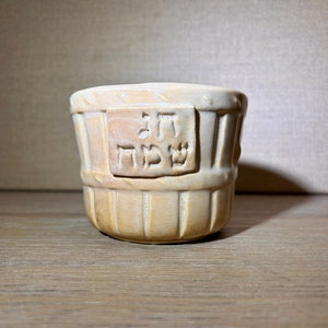 May include: A beige ceramic cup with a basket-weave texture and a rectangular plaque on the front featuring the Hebrew text "חג שמח" embossed in the clay. The vessel has a rounded shape with vertical indentations and a smooth, matte finish.