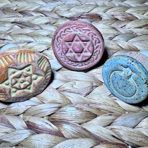 May include: Three decorative bottle stoppers with ceramic tops. One is orange with a star design, another is pink with a Star of David, and the third is blue with a heart. The stoppers are set against a woven background.