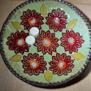 May include: A green ceramic plate with a decorative floral design in shades of red, orange, and blue. The plate has a scalloped edge and the word "Earth" is written on the side. Two white tea lights are placed on the plate.