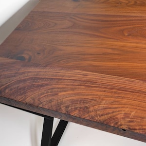 May include: Close-up of a wooden table with a rich, warm brown color. The table has a natural wood grain pattern and is supported by black metal legs. The table's surface is smooth and appears to be well-finished.