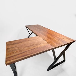 May include: A wooden L-shaped desk with a natural wood grain finish. The desk has a rectangular top and is supported by black metal legs. The desk is designed for a corner space and is suitable for a home office or workspace.