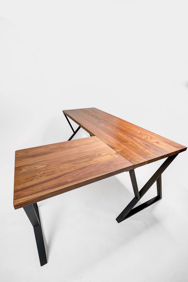 May include: A wooden L-shaped desk with a natural wood grain finish. The desk has a rectangular top and is supported by black metal legs. The desk is designed for a corner space and is suitable for a home office or workspace.