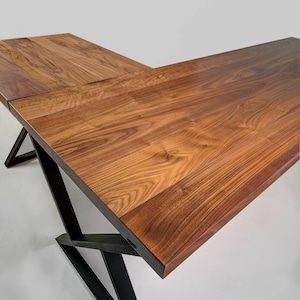 May include: A large, L-shaped wooden desk with a rich, brown finish. The desk features a modern design with black metal legs that provide support. The wood grain is visible, adding to the desk's natural aesthetic.