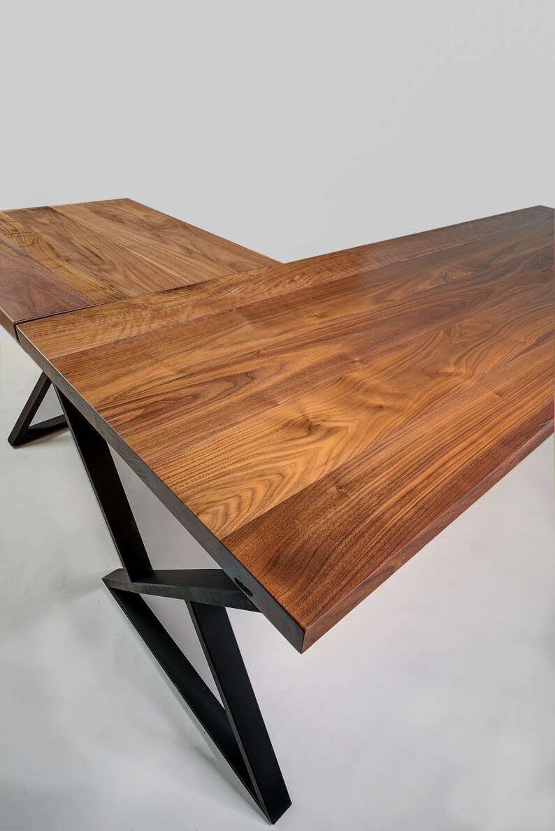 May include: A large, L-shaped wooden desk with a rich, brown finish. The desk features a modern design with black metal legs that provide support. The wood grain is visible, adding to the desk's natural aesthetic.