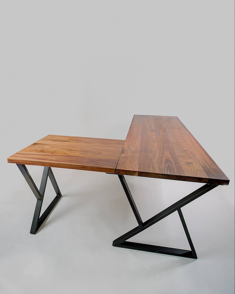 May include: A wooden desk with a natural finish and a unique L-shape design. The desk features a dark metal frame with angled legs. The desk is made of two connected rectangular surfaces.
