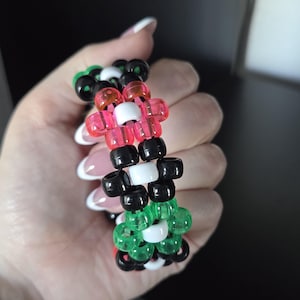 Può includere: Un braccialetto a più file realizzato con perline cilindriche in plastica. Il design presenta un motivo ripetuto di perline nere, bianche, rosa traslucide e verdi traslucide disposte in una struttura a griglia interconnessa.