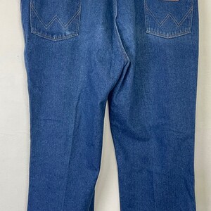 Może przedstawiać: Dżinsy z niebieskiego denimu z brązową sk&oacute;rzaną naszywką na tylnej kieszeni z napisem "Wrangler".