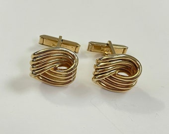 Puede incluir: Un par de gemelos de oro con un diseño de nudo. Los gemelos están hechos de un metal liso y brillante.