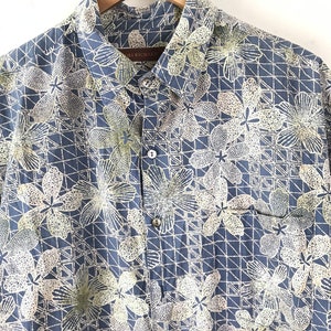 Puede incluir: Una camisa azul de botones con un estampado floral blanco y amarillo. La camisa tiene un bolsillo en el lado izquierdo y un cuello abotonado.