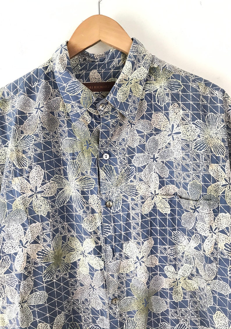Puede incluir: Una camisa azul de botones con un estampado floral blanco y amarillo. La camisa tiene un bolsillo en el lado izquierdo y un cuello abotonado.