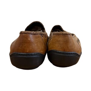 May include: A pair of brown leather slip-on shoes. The shoes have a dark brown sole and a stitched design on the back. The interior lining is a patterned fabric.