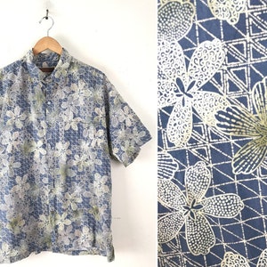 Puede incluir: Una camisa azul de manga corta con botones y un estampado floral blanco y verde. La camisa tiene un patr&oacute;n geom&eacute;trico en el fondo.