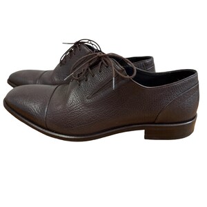 Peut inclure: Une paire de chaussures richelieu en cuir marron foncé avec une finition texturée et grainée. Ces chaussures présentent un bout droit, un petit talon et des lacets marron foncé assortis passant par des œillets sur le dessus de la tige.