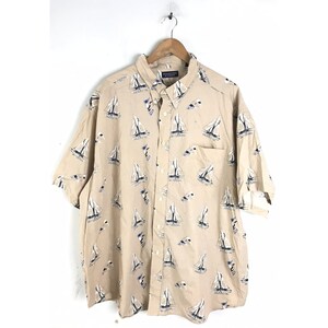 May include: A beige short-sleeved button-down shirt with a sailboat print. The shirt has a pocket on the left side.