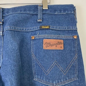 Może przedstawiać: Dżinsy z niebieskiego denimu z tylną kieszenią ozdobioną brązowym sk&oacute;rzanym naszywką z napisem "Wrangler" białymi literami.