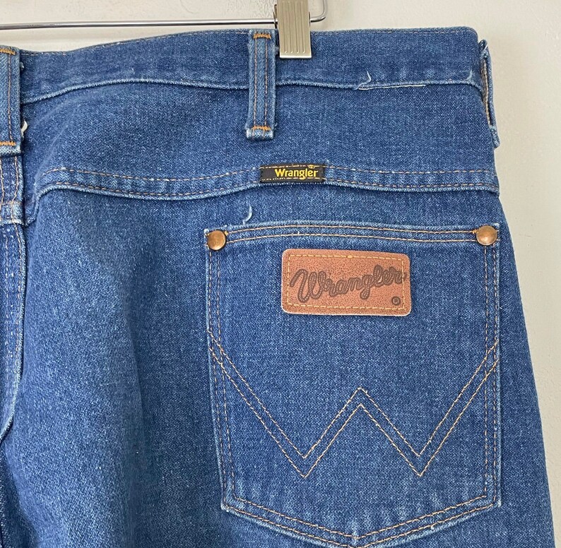Może przedstawiać: Dżinsy z niebieskiego denimu z tylną kieszenią ozdobioną brązowym sk&oacute;rzanym naszywką z napisem "Wrangler" białymi literami.