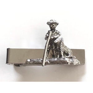 May include: Silver tie clip featuring a miniature figurine of a person with a dog. The person is holding a stick and wearing a hat. The figurine is mounted on a silver bar.