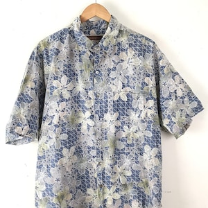 Puede incluir: Una camisa azul de manga corta con botones y un estampado floral blanco y verde. La camisa tiene un bolsillo en el lado izquierdo.