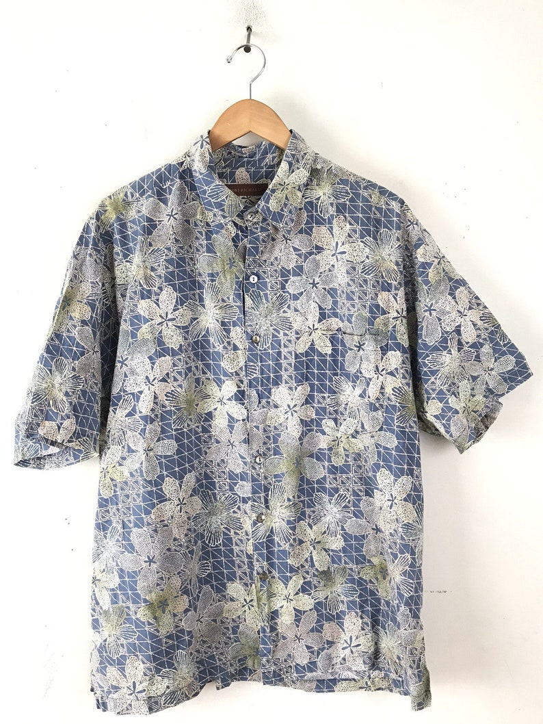 Puede incluir: Una camisa azul de manga corta con botones y un estampado floral blanco y verde. La camisa tiene un bolsillo en el lado izquierdo.