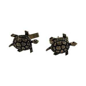 May include: A pair of cufflinks featuring a turtle design. The cufflinks are made of a metallic material with a detailed turtle shell pattern. The turtle's head, legs, and tail are also visible. The cufflinks are set against a white background.