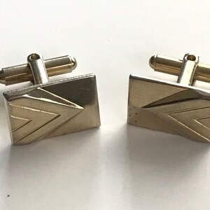 May include: A pair of gold-toned cufflinks with a geometric design. The cufflinks have a square shape with a pointed, triangular pattern.