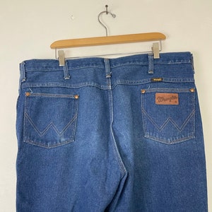 Może przedstawiać: Dżinsy z niebieskiego denimu z brązową sk&oacute;rzaną naszywką Wrangler na tylnej kieszeni.