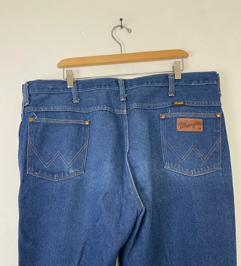 Może przedstawiać: Dżinsy z niebieskiego denimu z brązową sk&oacute;rzaną naszywką Wrangler na tylnej kieszeni.
