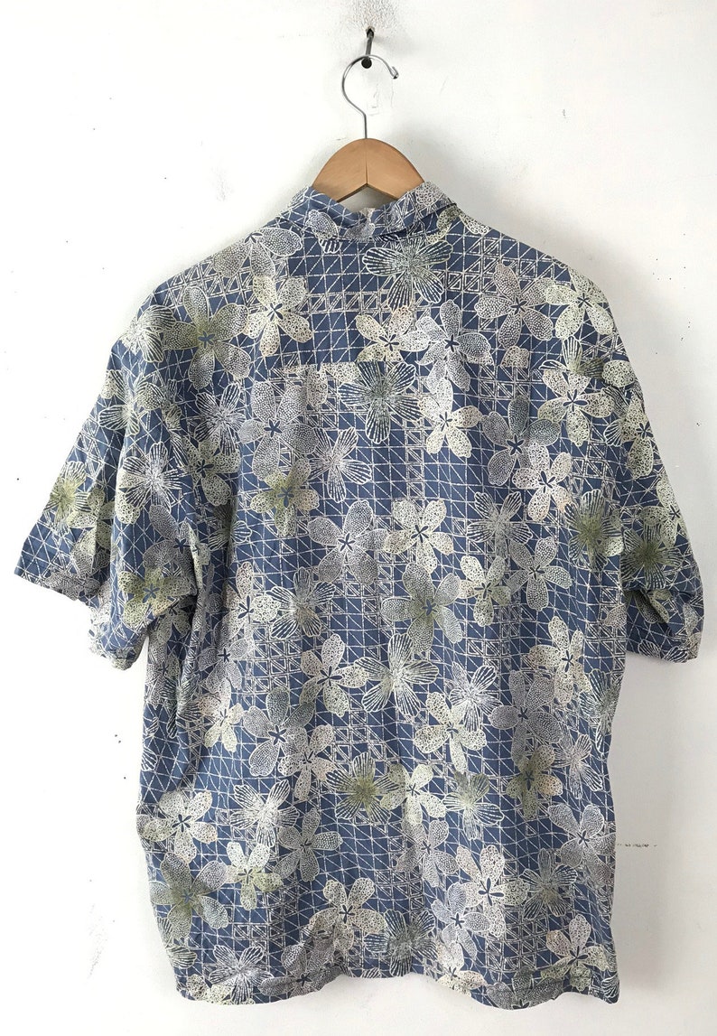 Puede incluir: Una camisa azul de manga corta con un estampado floral blanco y verde. La camisa tiene un cuello abotonado y un corte holgado.