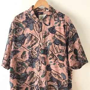 May include: A pink short-sleeved button-down shirt with a fishing-themed print. The print features fish, fishing lures, and other fishing-related imagery. The shirt has a button-down collar and a pocket on the left side.