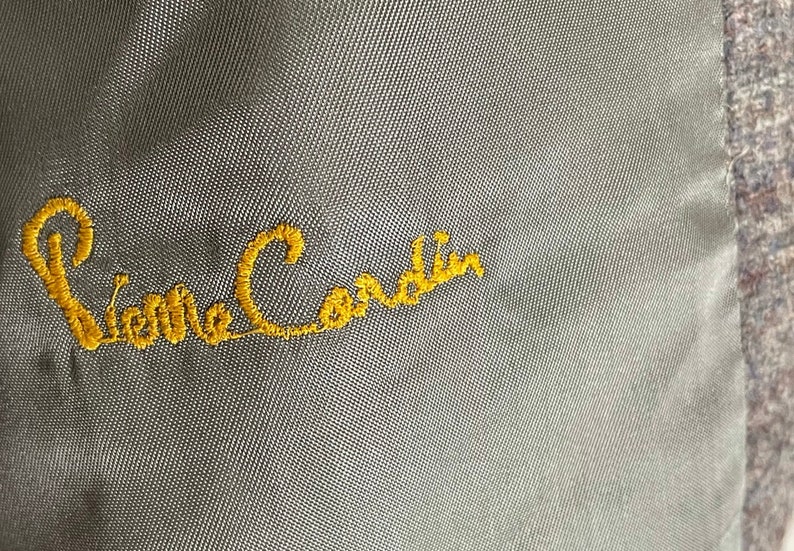 May include: A close-up of a gray fabric with the embroidered text "Pierre Cardin" in yellow thread.