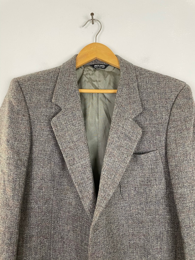 May include: A gray tweed blazer with a single button closure and a pocket on the left side. The label on the inside of the blazer reads "Pierre Cardin, Made in Hong Kong".