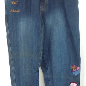 May include: Blue denim jeans featuring a faded wash, a button and zipper fly, and belt loops. The right thigh has embroidered text reading "By D" and "Meathook". The left leg features four circular patches with "Harlem" and "H" branding.