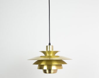 May include: A gold-colored pendant light fixture with a tiered, layered design. The light hangs from a black cord against a white background. The fixture has a series of overlapping, circular shades, creating a unique, modern aesthetic.