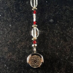 Może przedstawiać: Srebrny charm z geometrycznym wzorem wisi na koralikowej bransoletce z przezroczystymi i czerwonymi koralikami. Białe pióro jest przymocowane do górnej części bransoletki.