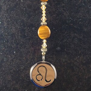 May include: A gold and brown beaded car charm with a tiger's eye stone and a silver charm with the Leo zodiac symbol.