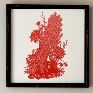 May include: A square black frame containing a white background with a red paper-cut style illustration of a peacock perched on a rock. The peacock is surrounded by detailed floral elements and foliage, creating a symmetrical and intricate design.