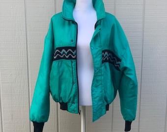 May include: A teal blue vintage bomber jacket with a black zipper and snap closure. The jacket has a black and white geometric pattern on the chest and sleeves.