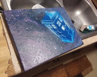 May include: A blue and white TARDIS, a time machine from the Doctor Who television series, is painted on a canvas with a galaxy background.