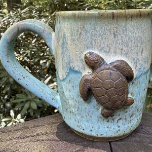 May include: A ceramic mug with a speckled light blue and cream glaze featuring a raised, brown, textured sea turtle sculpture on the front. The mug has a large, curved handle and a cylindrical body with a smooth rim and a sturdy base.