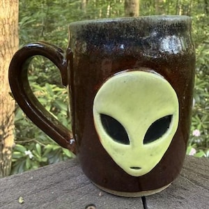 May include: A dark brown ceramic mug featuring a raised, light green alien face appliqué on the front. The mug has a glossy finish, a sturdy handle on the left side, and a dark, mottled rim. The alien design includes large, smooth, black eyes.