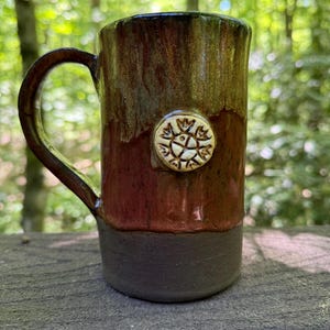 May include: A tall ceramic mug with a handle, featuring a two-tone glaze of dark brown at the base and a blend of reddish-brown and olive green on the upper portion. A small, circular, cream-colored medallion with a stamped floral design is centered.