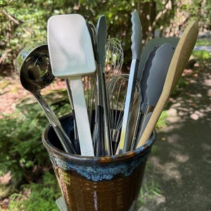 May include: A ceramic utensil holder filled with various kitchen tools. The tools include a stainless steel ladle, whisks, spatulas, tongs, and a wooden spoon. The holder is brown with a blue rim, and the tools are arranged in a vertical display.