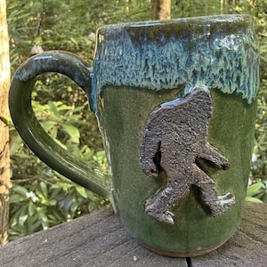 May include: A ceramic mug with a glossy green glaze and a dark blue drip pattern around the rim. A textured, dark brown, raised relief figure of a bipedal creature is attached to the side of the mug, featuring a curved handle on the left side.