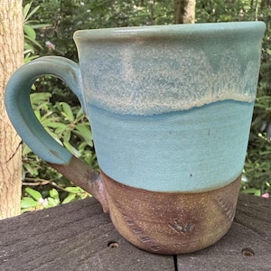 May include: A ceramic mug featuring a two-tone glaze design. The upper portion is a light blue with a textured, speckled finish, while the lower section and the curved handle are a solid, earthy brown with subtle, diagonal impressed markings.