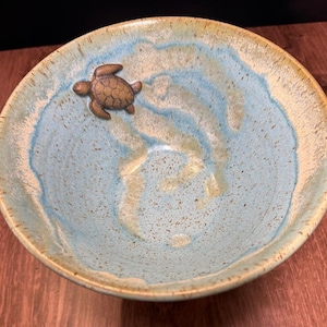 May include: Ceramic bowl with a light blue and beige glaze, featuring a bronze-colored turtle figurine on the interior. The bowl has a wide rim and a speckled texture, evoking a coastal theme.