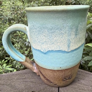 May include: A ceramic mug featuring a three-tone glaze with a textured brown base, a vibrant turquoise middle section, and a pale mint green upper rim. The mug has a sturdy, curved handle that transitions from brown to turquoise, with a smooth finish.